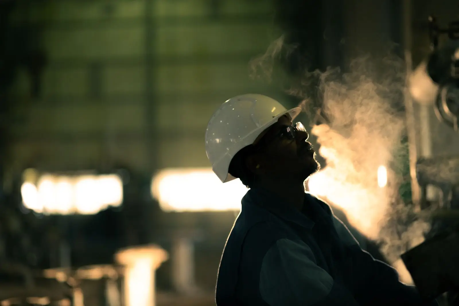A plant operator in hard hat and safety glasses beside a fired boiler, steam rising against the furnace glow
